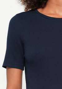 Γυναίκα που φοράει μια εφαρμοστή, κοντομάνικη μπλε ριμπ πουλόβερ που αναδεικνύει την υφή και την λείο στρογγυλό λαιμό.