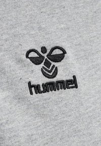 Tessuto grigio con un logo ricamato di colore nero a forma di ape sopra la parola "hummel" in un carattere audace. Texture liscia, design informale.