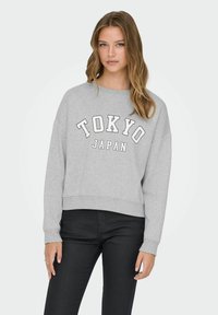 Ung kvinde med langt hår iført en lysegrå sweatshirt med teksten "TOKYO JAPAN" og sorte bukser, stående med en neutral udtryk.