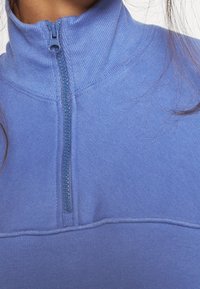 Polaire bleu à fermeture éclair avec un col haut, présentant un tissu texturé, une fermeture éclair centrale et des accents côtelés le long du col et de l'ourlet.