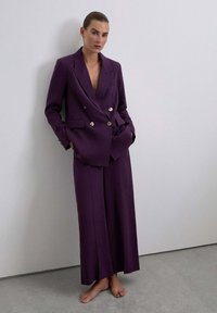 Blazer púrpura de doble botonadura con botones dorados, combinado con pantalones de pierna ancha. Confeccionado en un material suave, presenta un escote en V profundo.