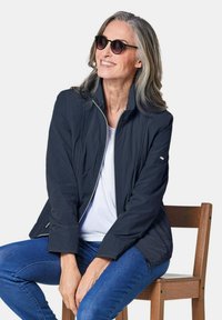 Marineblaue leichte Jacke mit Reißverschluss, vertikalen Nähten und seitlichen Taschen. Über einem weißen Hemd getragen, kombiniert mit blauen Jeans und Sonnenbrille.