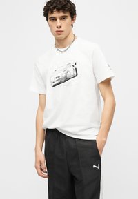 Ung mann iført en hvit T-skjorte med et bilmotiv og svarte Puma-bukser, poser med den ene hånden i lommen mot en hvit bakgrunn.