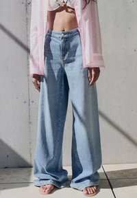 Hellblaue weite Jeans mit hoher Taille, ausgestattet mit einem Knopfverschluss und einem lockeren Schnitt. Getragen mit einem kurzen weißen Oberteil und einem rosa gestreiften Hemd.