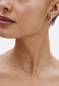 Srebrny naszyjnik z łańcuszkiem z małymi okrągłymi koralikami oraz dopasowana kolczyk kołowy zdobiony przezroczystymi kamieniami szlachetnymi. Odcień skóry jest naturalny.