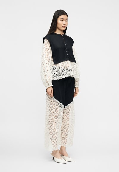 Γυναίκα που στέκεται φορώντας μια λευκή δαντελένια μπλούζα και παντελόνια με φαρδιές βάσεις με μαύρα υφασμάτινα πάνελ, σε συνδυασμό με λευκές μυτερές μύτες με χαμηλά τακούνια.