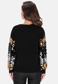 Schwarzer Pullover mit langen Ärmeln und großen schwarzen, goldenen und silbernen Pailletten. Rippbündchen an Ärmeln und Saum, weiche Stoffstruktur.