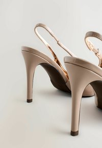 Stilettos de charol en tono nude con punta cerrada, correa ajustable en el talón, detalles metálicos y tacón alto y delgado. Textura suave.