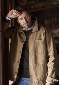 Braune Baumwolljacke mit Kragen und Knopfverschluss, ausgestattet mit einer Brusttasche und einer strukturierten Oberfläche, über einem dunklen Pullover getragen.