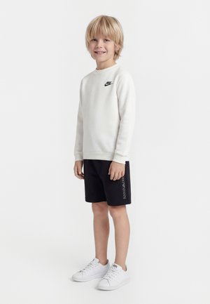 Lyshåret dreng smilende, iført hvid Nike-sweatshirt, sorte Emporio Armani-shorts og hvide sneakers, stående foran en ensfarvet hvid baggrund.