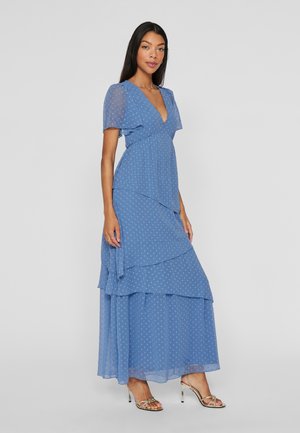 Femme portant une robe maxi bleue à volants avec des manches courtes transparentes et des pois, associée à des sandales à talons hauts dorées à lanières.