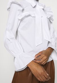 Blusa bianca con bordi arricciati e collana a fiocco. Presenta maniche lunghe con polsini a campana. Tessuto liscio con una consistenza croccante.