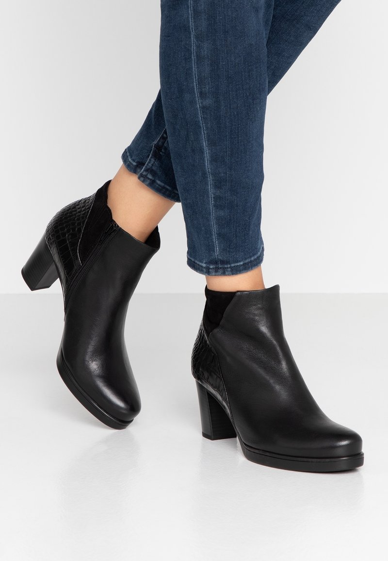 Bottes mi-mollet en cuir noir à talons carrés portées avec un jean bleu foncé court sur un fond blanc uni.
