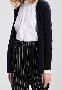 Mörk marinblå cardigan över vit skjorta med krage, kombinerad med svarta och vita randiga byxor. Har en avslappnad passform och ribbat detaljer.