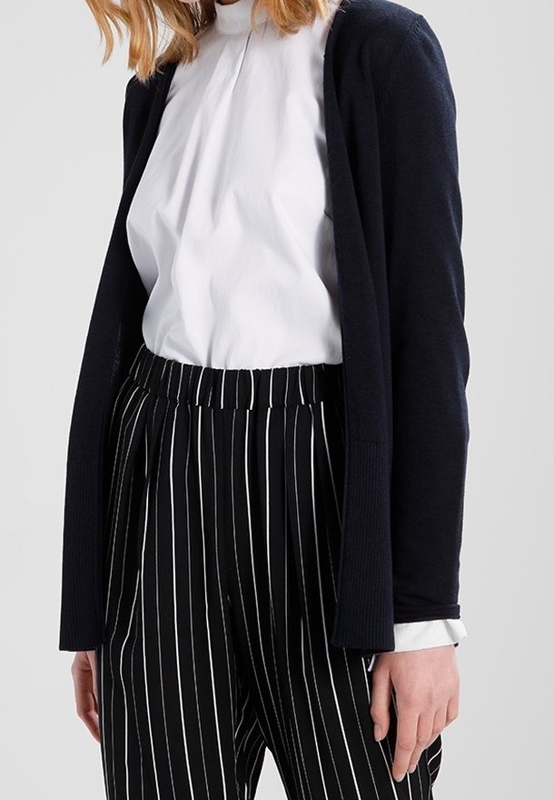 Mörk marinblå cardigan över vit skjorta med krage, kombinerad med svarta och vita randiga byxor. Har en avslappnad passform och ribbat detaljer.