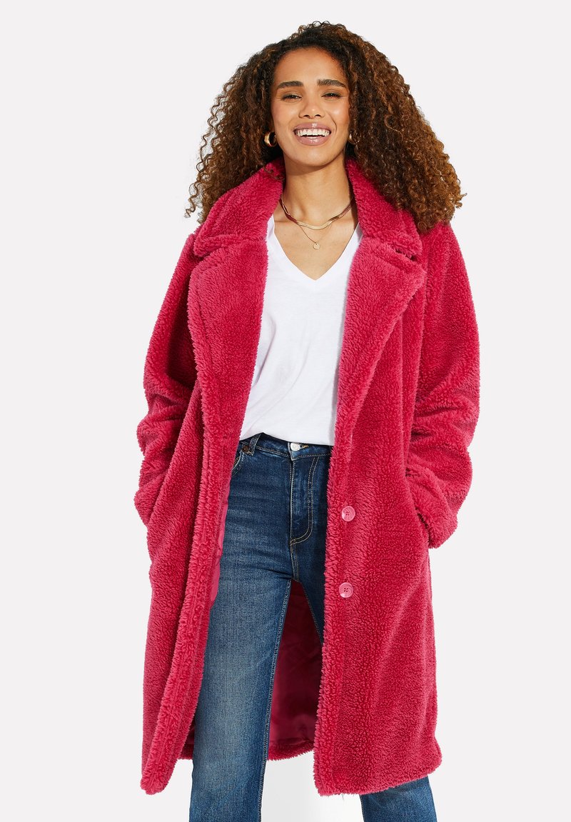 Luddig rosa lång kappa med krage, framknappar och sidofickor, bärs över en vit V-ringad tröja och blå jeans.