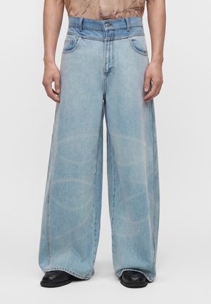 Άνδρας που φοράει ανοιχτό μπλε τζιν παντελόνια με φαρδιά μπατζάκια και μαύρα παπούτσια, στέκεται μπροστά σε ένα απλό λευκό φόντο.