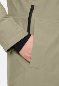 Hand gedeeltelijk in een beige jaszak met zichtbare zwarte manchet en zwart verticale ritsdetail nabij de zak.