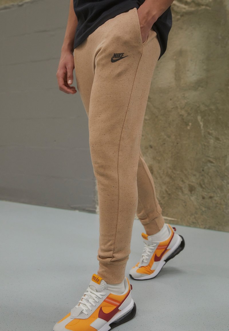 Beige Nike sweatpants med en svart logotyp, avsmalnande passform och sidofickor. Matchas med färgglada Nike-sneakers som har orange och röda inslag.