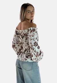 Blusa off-the-shoulder bianca con motivo floreale marrone, dotata di maniche a sbuffo e vita arricciata; abbinata a jeans in denim blu chiaro.