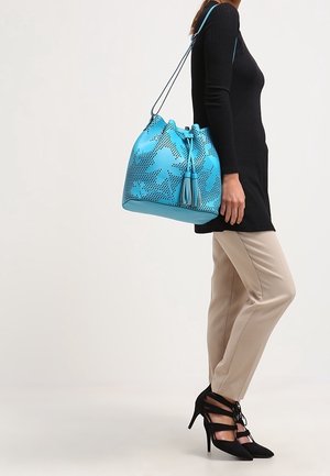 Donna che indossa pantaloni beige e un top nero porta una grande borsa blu con design perforato e dettagli con frange sulla spalla.