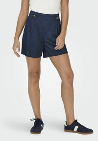 Shorts bleu marine à taille haute, avec des plis et des boutons dorés ; fabriqués dans un tissu doux. Associés à des baskets bleu marine avec des détails blancs.
