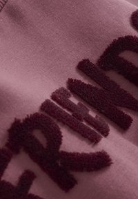 Polarowa bluza w stonowanym różu z wyraźnym bordowym napisem. Miękka tekstura, dopasowany krój i widoczne detale szwów.