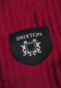 Pachet cu logo Brixton pe material matlasat roșu; design cu scut negru ce prezintă text alb și motive stilizate de lei; suprafață texturată.