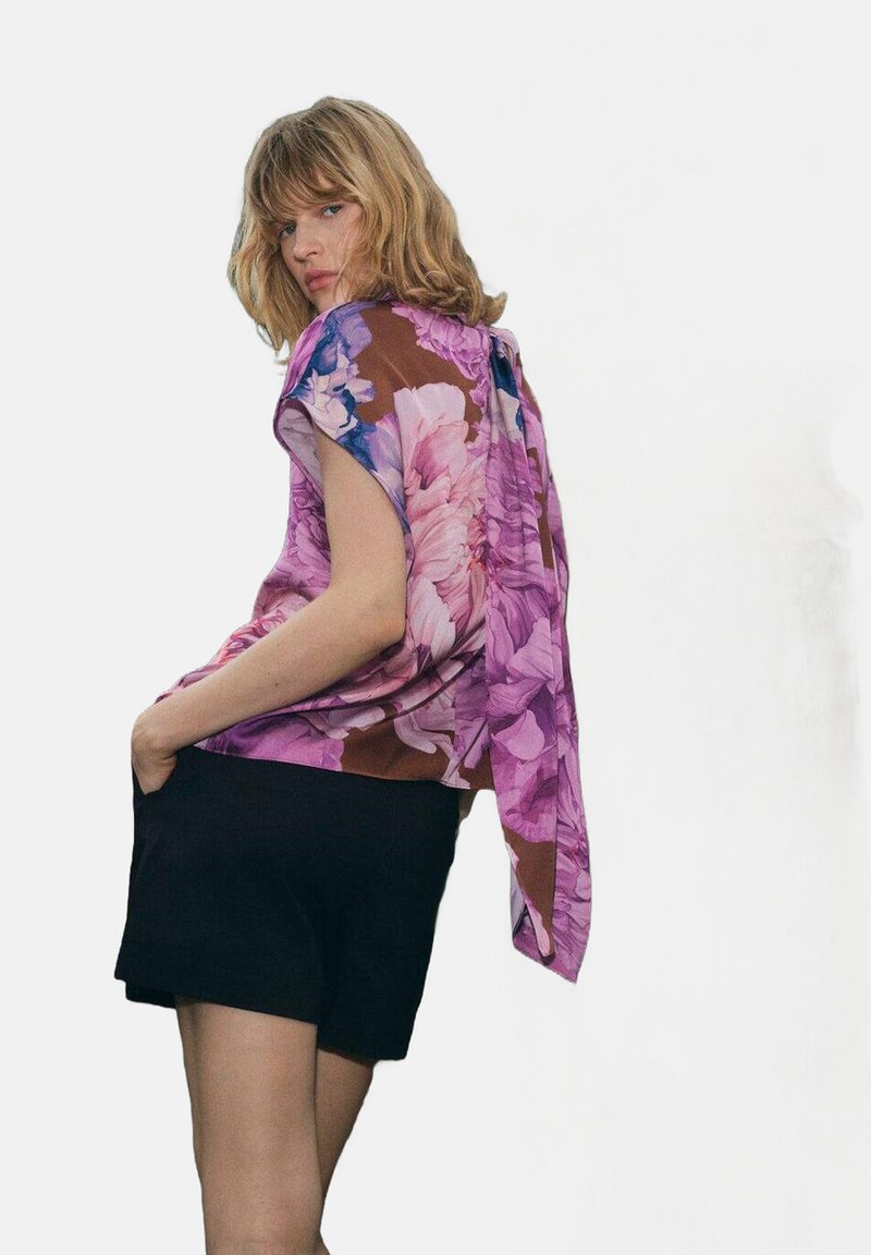 Femme blonde portant un blouse à fleurs violette à manches courtes avec un short noir ample, posant les mains dans les poches devant un fond uni.
