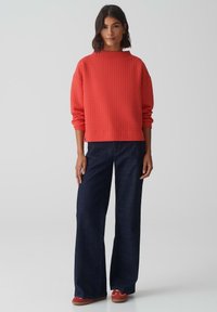 Pull rouge côtelé avec un col haut, porté avec un pantalon en denim noir à jambes larges et des chaussures rouges, présente des manches longues et une coupe décontractée.