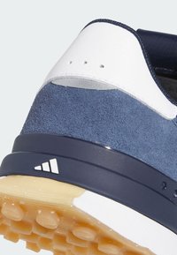 Tallone di una sneaker in pelle scamosciata blu navy e pelle bianca con superficie testurizzata, caratterizzato da una intersuola in gomma blu scuro e una suola esterna in gomma color marrone chiaro.