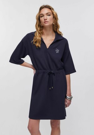 Femme aux cheveux blonds ondulés portant une robe bleu marine longueur genou avec manches retroussées, col en V, taille cintrée par un cordon et bracelets argentés.