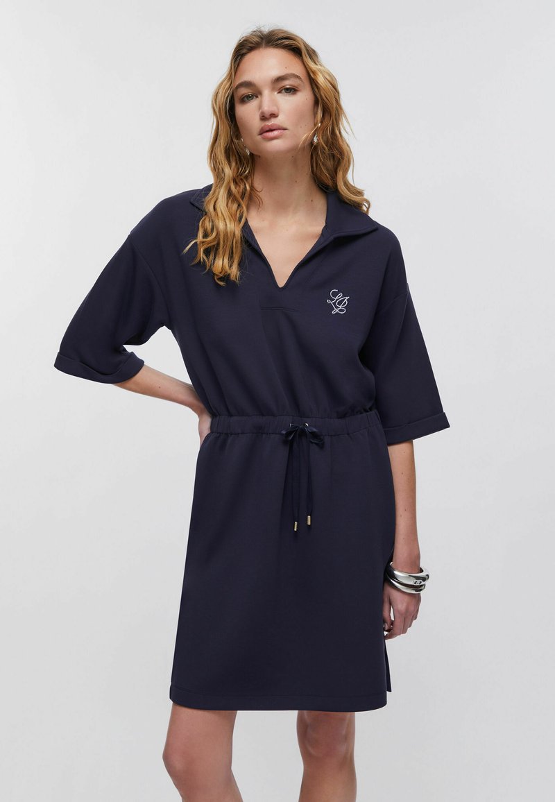 Mujer con cabello rubio ondulado que lleva un vestido azul marino hasta la rodilla con mangas remangadas, cuello en V, cintura con cordón y pulseras plateadas.