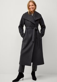 Cappotto in lana carbone con ampio colletto, vita con cintura e tasche laterali; indossato sopra pantaloni in pelle nera e stivaletti neri a punta.