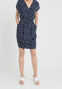 Dunkelblaues Kleid mit weißen Punkten, V-Ausschnitt, kurzen Ärmeln, elastischem Bund und seitlichen Taschen. Kombiniert mit weißen Blockabsatzsandalen.