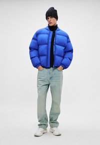 Mėlynas pūkų švarkas su siuvinėtu dizainu, užtrauktuku, o po juo juoda polo vilna, derinama su šviesiai mėlynomis džinsomis ir baltais sportbačiais.