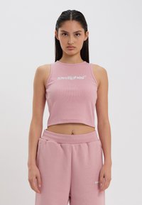 Top scurt roz cu nervuri și logo alb, având un guler înalt și un design fără mâneci. Stilizat cu pantaloni sport roz asortați.