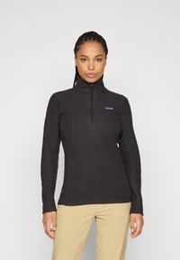 Donna con capelli intrecciati che indossa un fleece nero con zip e pantaloni beige, in piedi contro uno sfondo chiaro e semplice.