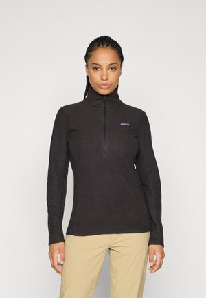 Vrouw met gevlochten haar, gekleed in een zwarte textuur rits-fleece en beige broek, staande tegen een egale lichte achtergrond.