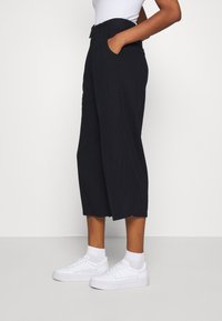 Svarta korta byxor med skräddarsydd design, midja med bältesöglor, sidofickor och slät textur, kombinerade med vita sneakers.