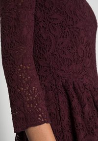 Burgunderröd spetsklänning med trekvartslånga ärmar, med intrikata blommönster, texturerad tyg och en utsvängd midja.