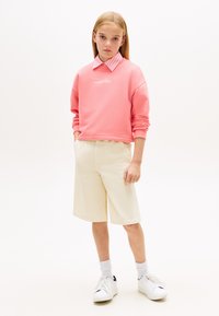 Ροζ φούτερ με λευκό λογότυπο, ριγέ πουκάμισο με γιακά από κάτω, μπεζ σορτς και λευκά αθλητικά παπούτσια με μαύρες λεπτομέρειες. Απλό φόντο.