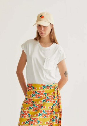 Mujer con gorra beige con bordado naranja, camiseta blanca con bolsillo y falda envolvente amarilla con patrones de frutas naranja y verdes.
