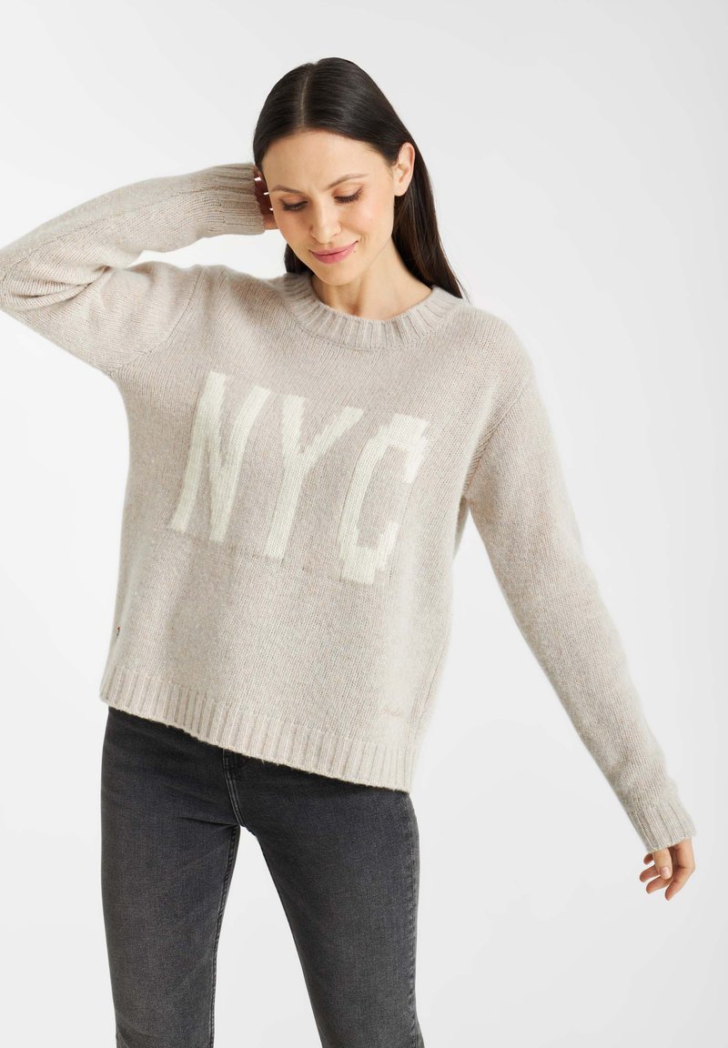 Lichtbeige gebreide trui met een groot wit "NYC"-grafiek. Geribde ronde halslijn en manchetten, met een relaxte pasvorm en zachte textuur.