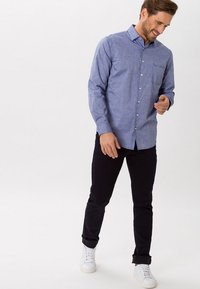 Camicia azzurro chiaro con bottoncini in un tessuto testurizzato, con taschino sul lato sinistro del petto, abbinata a pantaloni scuri e sneakers bianche.