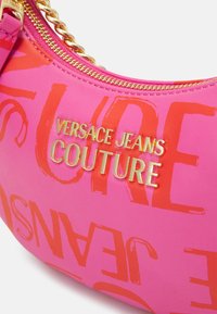 Bolso de hombro sintético rosa con texto naranja llamativo y logo dorado "VERSACE JEANS COUTURE". Cuenta con cremallera y adorno de cadena en tono dorado.