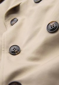 Beżowa tkanina o gładkiej fakturze z dużymi ciemnymi guzikami. Guziki są błyszczące, mają cztery otwory i wytłoczone napisy na obrzeżu.