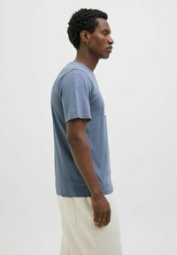 Heren crewneck t-shirt van gedempt blauwe jersey stof, met korte mouwen en een subtiele graphic print op de borst. Gepaarde met lichtgekleurde broeken.