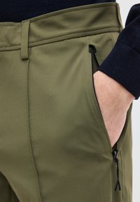 Χέρι στην τσέπη χακί πράσινου παντελονιού με εμφανή μαύρη τσέπη με φερμουάρ και μανίκι μαύρου πουλόβερ.