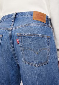 Jeans de mezclilla azules con un acabado texturizado, que presentan costuras de contraste, una etiqueta de marca de cuero y una etiqueta roja en el bolsillo trasero.